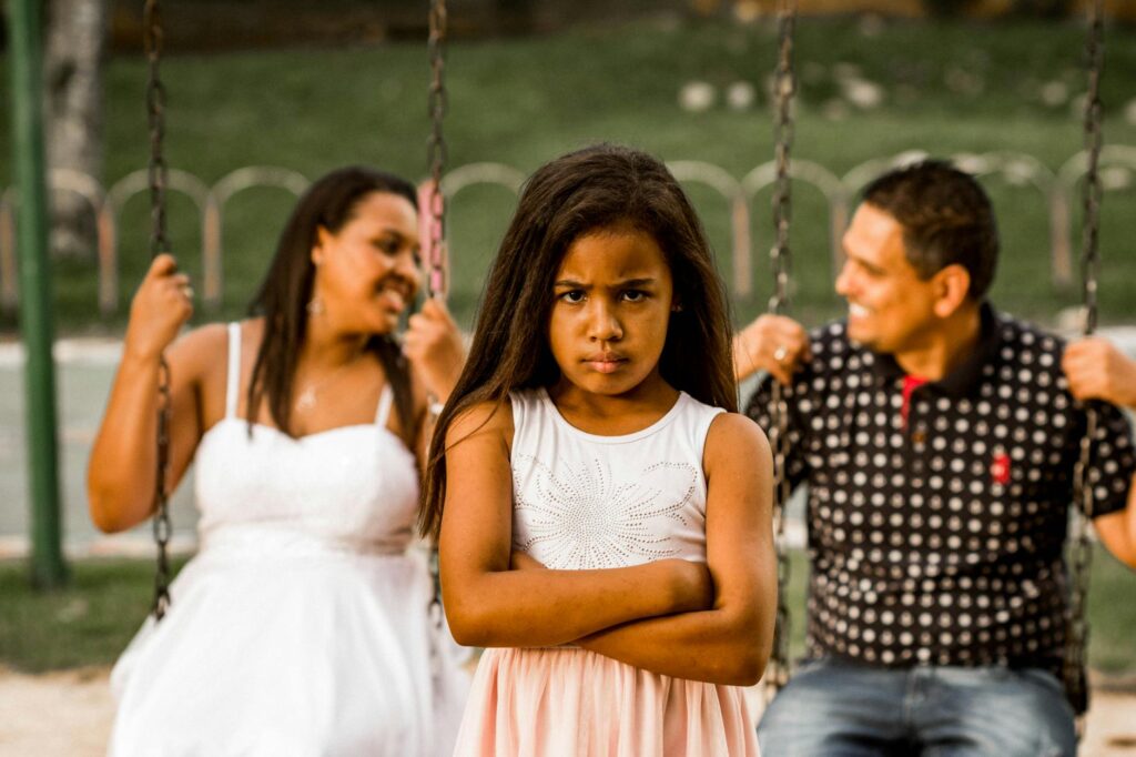 O que acontece quando a alienação parental é provada - Robison Souza - Perito em Psicologia Forense A young girl looking grumpy while her parents enjoy swings in a park.
